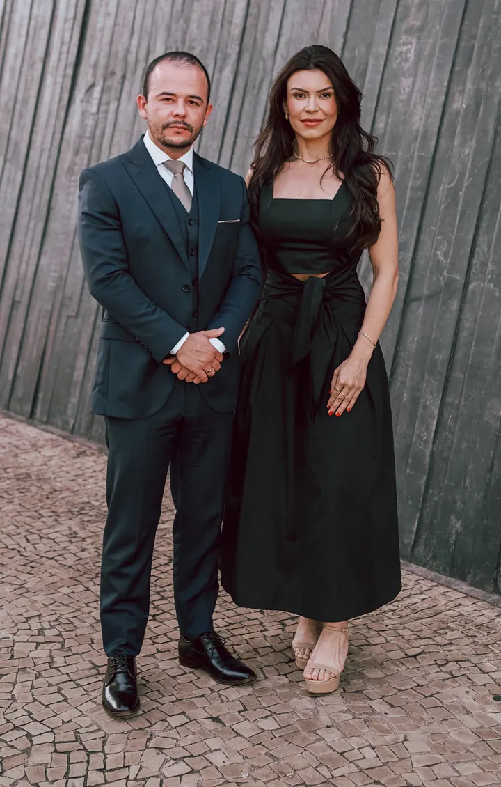 Dr. André de Sá e Dra. Emanuelle Jamille de corpo inteiro, diante de uma parede de madeira escura